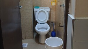 A small bathroom with a toilet, a bucket, and cleaning supplies on the wall. - Studio airbnb mirema drive (Nairobi)