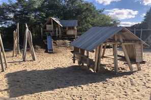 Ein Strandabschnitt mit Sand, Holzspielgeräten und einem kleinen Gebäude mit einer Metallbedachung.