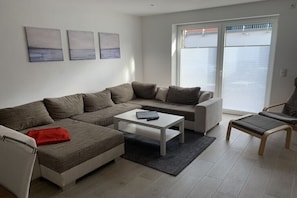 Ein modernes Wohnzimmer mit einem Ecksofa, einem kleinen Couchtisch und einem Sessel. An der Wand hängen drei Gemälde.