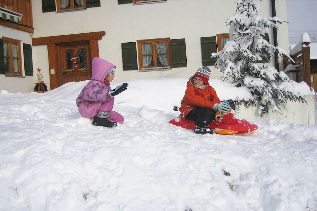 Two children sledding in a snowy outdoor area with a building in the background.