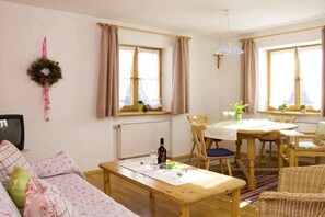 A cozy living room with a sofa, a wooden table, and a dining area with chairs and a window with curtains. - 4 - at the Füssen equestrian center (Weißensee-Füssen)