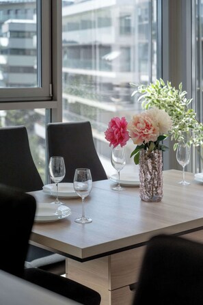 Ein Esstisch mit zwei Weingläsern, einer Blumenvase und Blick auf Gebäude durch große Fenster.
