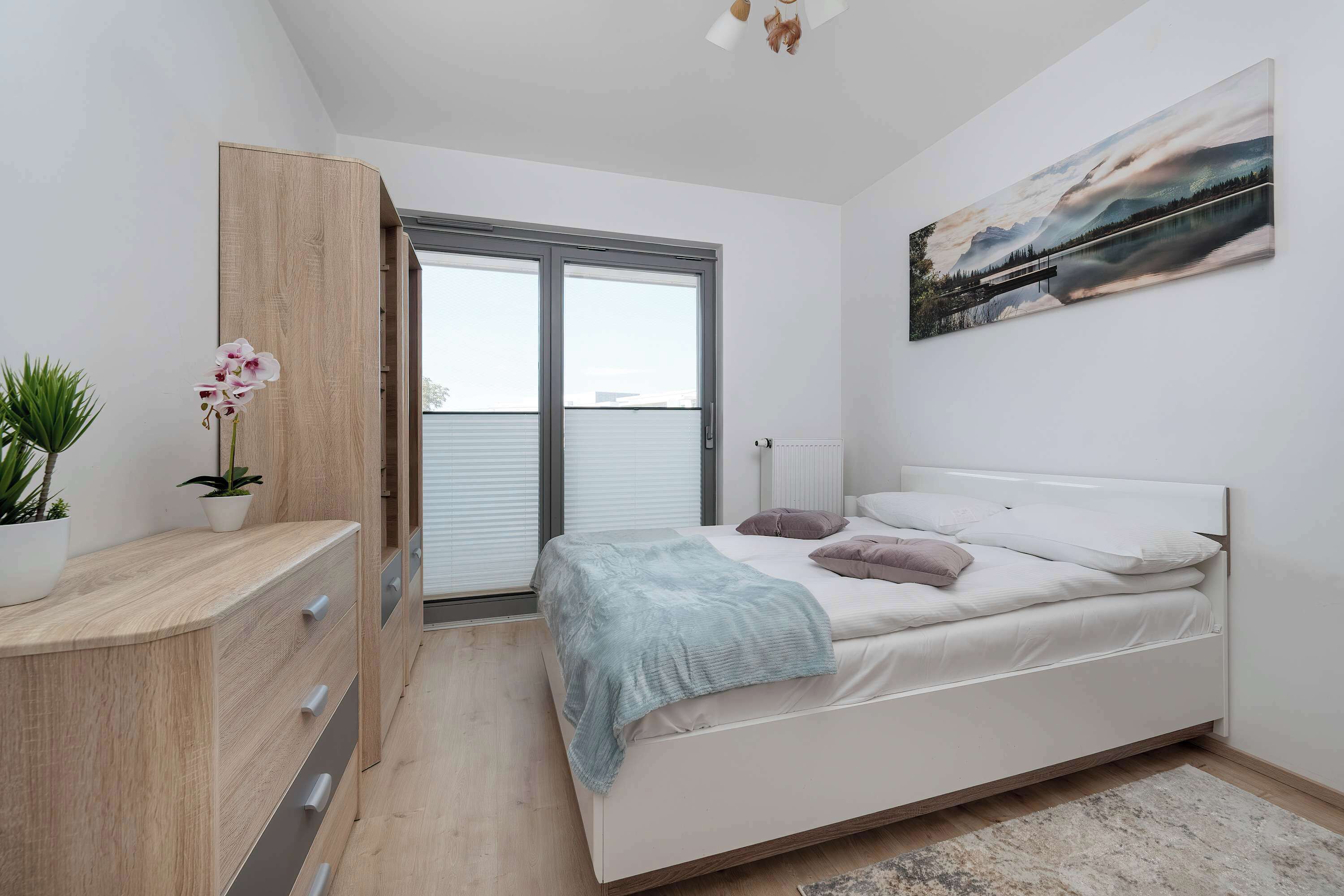 A bedroom with a bed, a nightstand, a window with blinds, a radiator, a painting on the wall, and a plant.