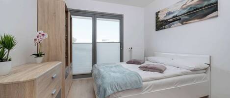 A bedroom with a bed, a nightstand, a window with blinds, a radiator, a painting on the wall, and a plant.