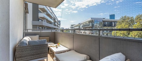 Un balcón con un sofá de mimbre, una mesita con un cuenco de fruta y vistas a un edificio moderno.