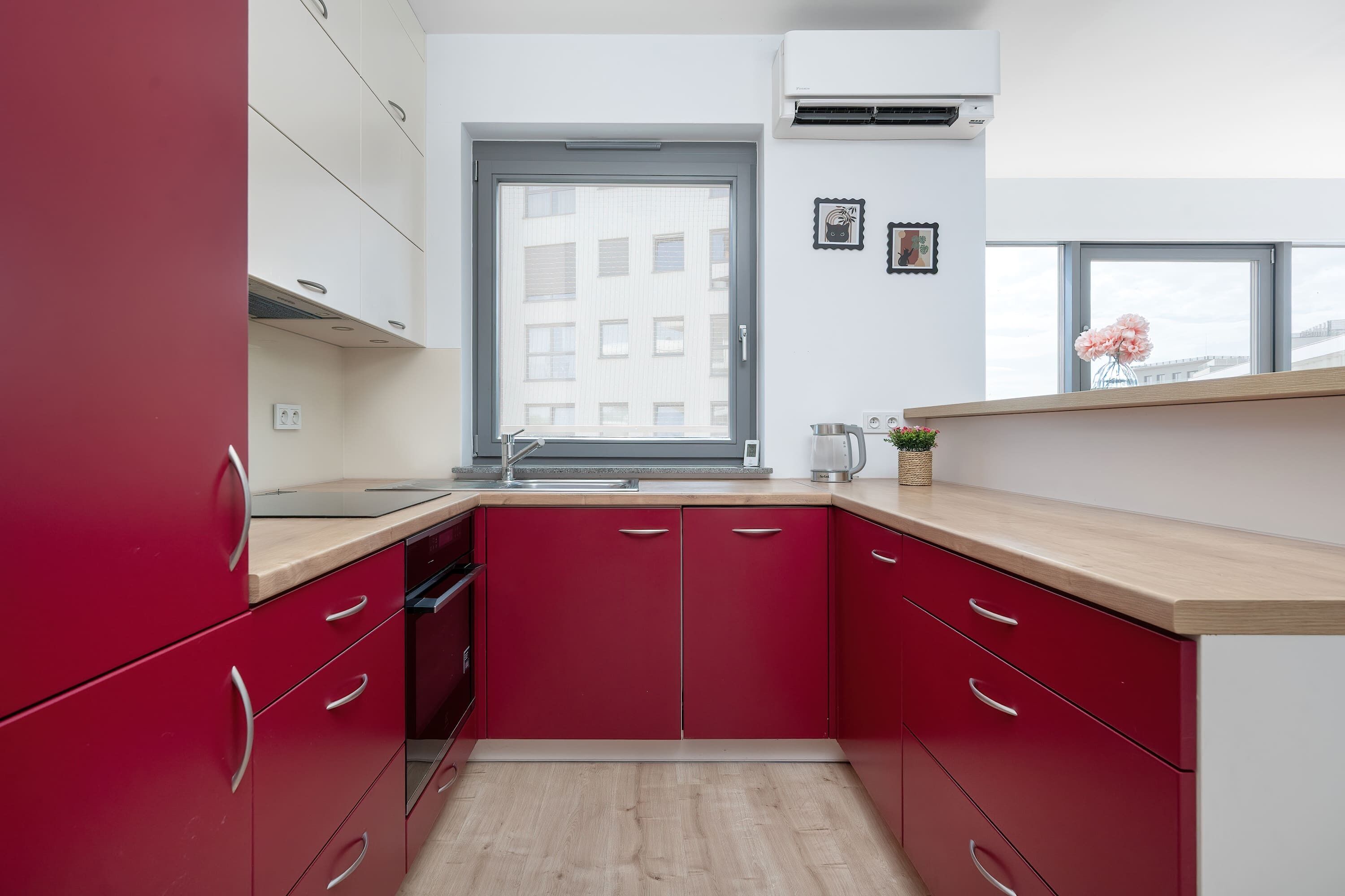 A compact kitchen with red cabinets, a wooden countertop, and a window with a city view.