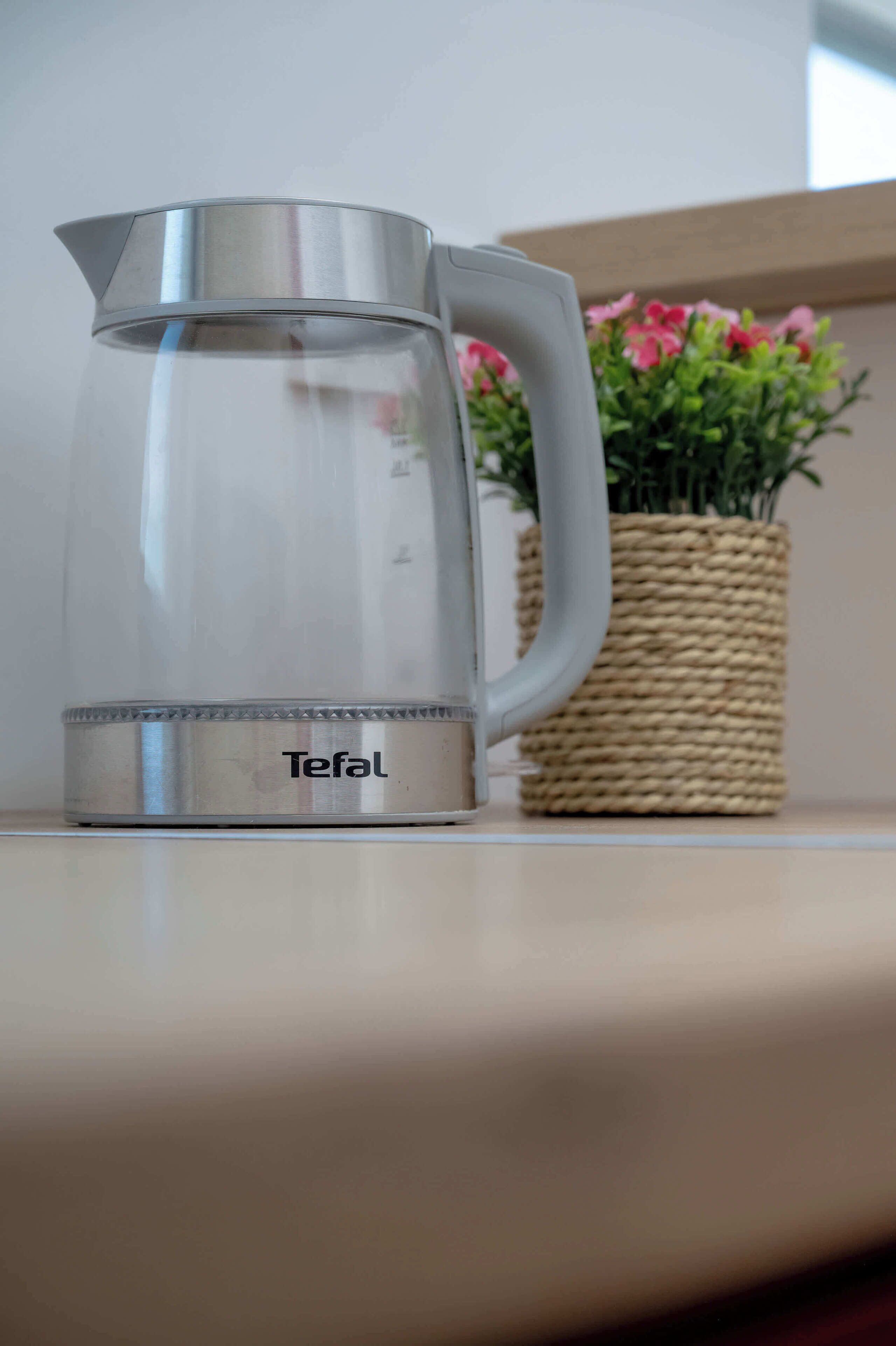Una teiera in acciaio inossidabile Tefal su uno scaffale, con un cesto intrecciato e fiori rosa sullo sfondo.