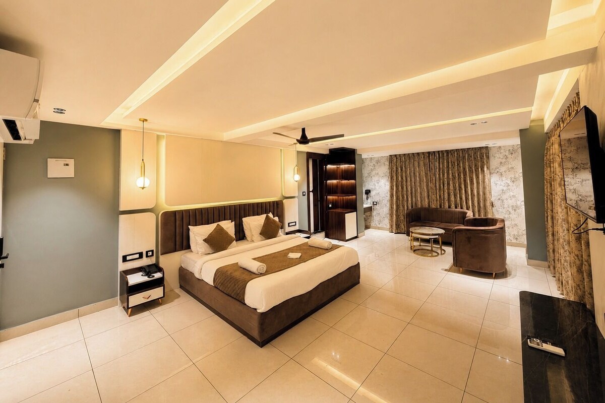 A modern hotel room with a large bed, a seating area, a TV, and a minibar.