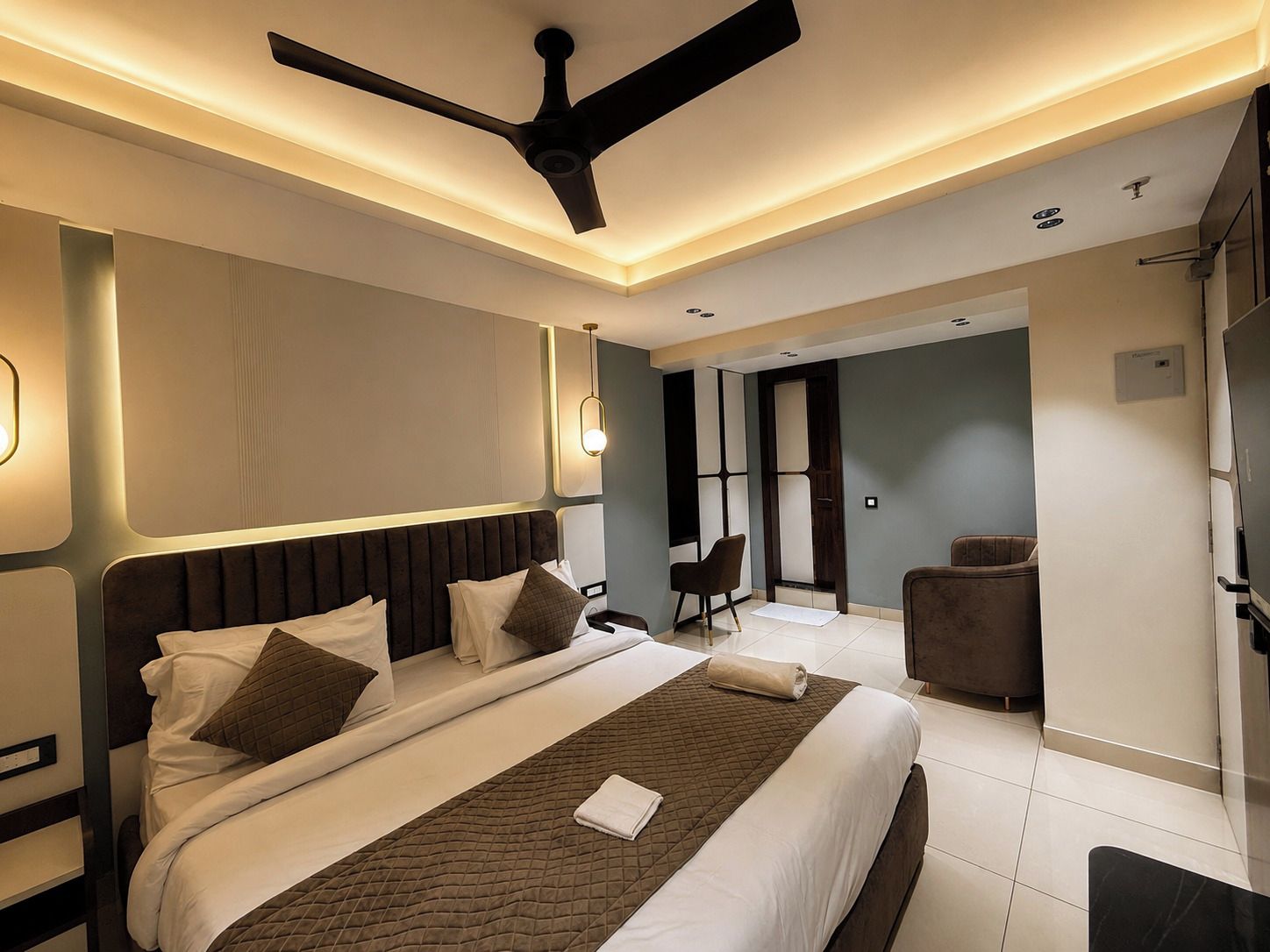 Quarto de hotel moderno com uma cama grande, um ventilador de teto e um espaço de estar com uma cadeira e uma mesa pequena.