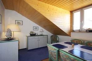 Ein Zimmer mit schrägverlegtem Holzboden, einem Esstisch mit Stühlen, einer kleinen Kommode und einem Fenster.