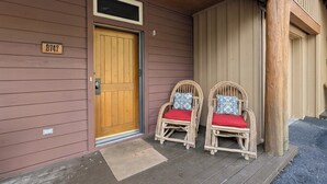 En dörr av trä med en nummerplatta, två flätade stolar med röda kuddar och en veranda med ett träräcke.