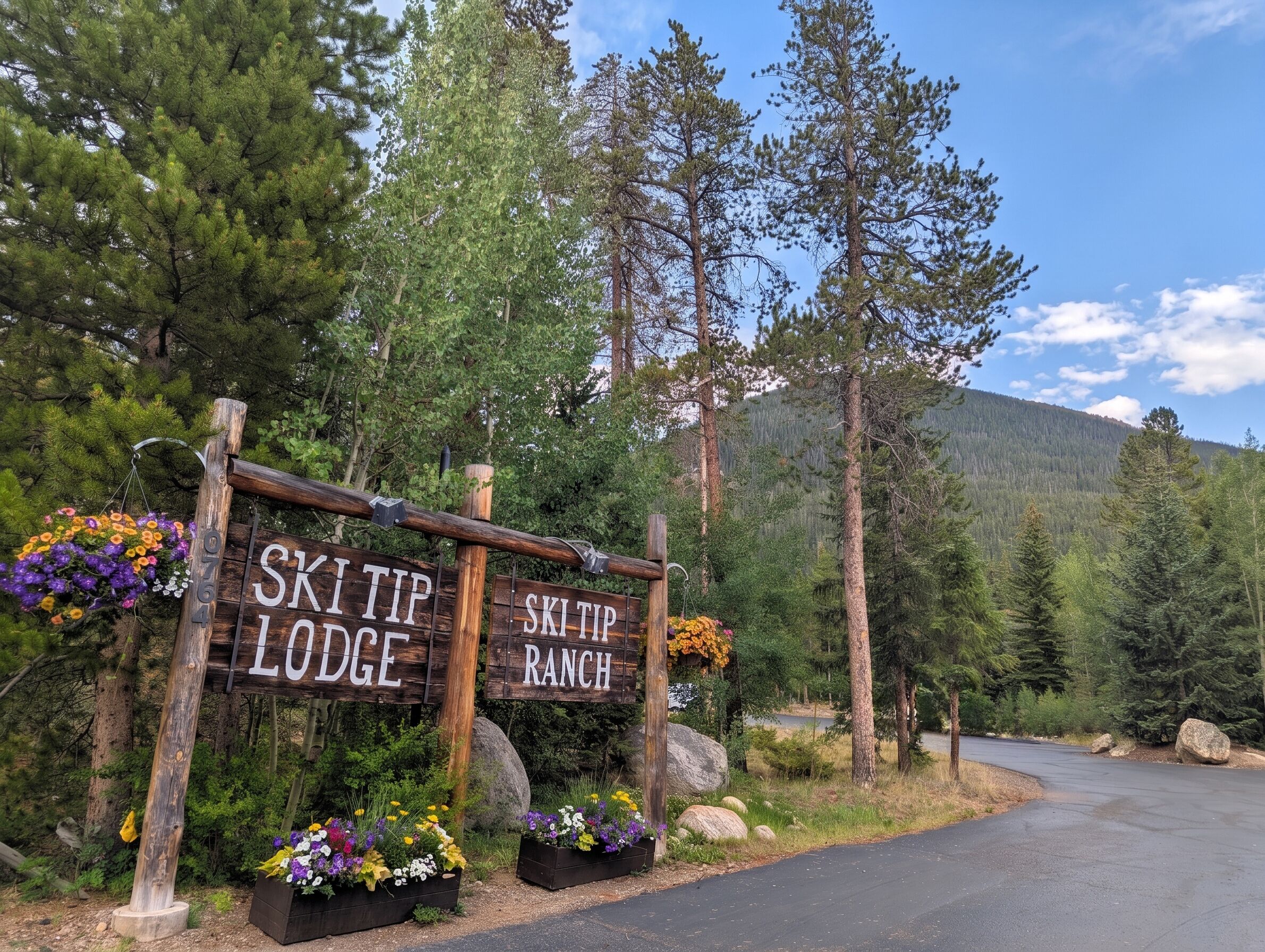 Un letrero de madera con la inscripción 'SKITIP LODGE' y 'SKI TIP RANCH', rodeado de follaje y una montaña al fondo.