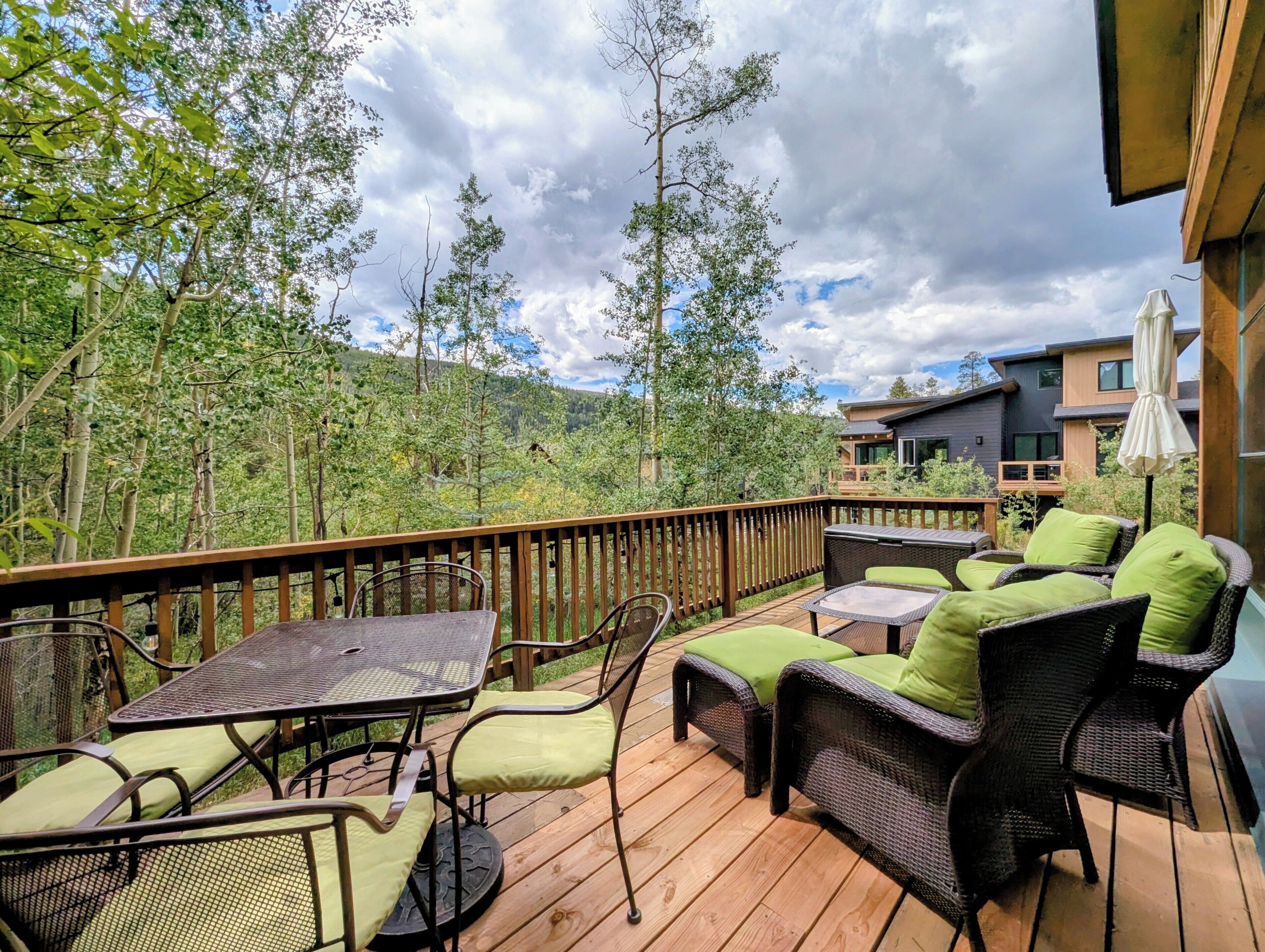 Una terraza de madera con mobiliario exterior, incluyendo una mesa y sillas, con vista a una zona boscosa y un edificio moderno al fondo.