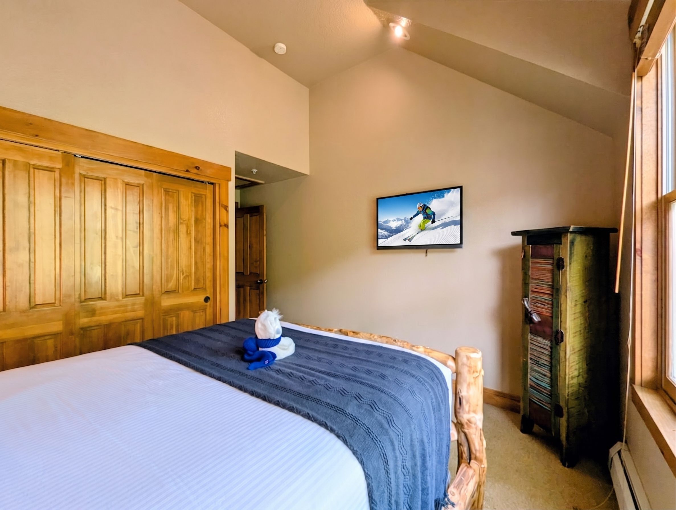 Un dormitorio con una cama, un armario de madera, un televisor montado en la pared y una mesita.