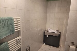 Un bagno con pavimento piastrellato, un portascope con un asciugamano verde, un termosifone bianco e un piccolo angolo lavabo con un asciugamano e un panno.