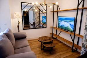 En moderne stue med en sofa, et lille rundt bord, et fjernsyn monteret på en træhylle og et spejl med geometrisk ramme.