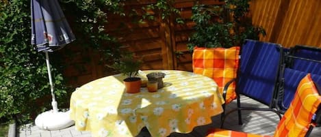 A round table with a floral tablecloth, two chairs with orange and blue cushions, and a potted plant on the table.