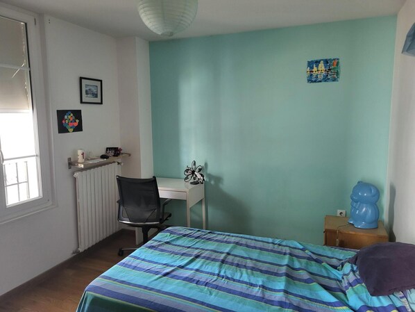 A bedroom with a bed, a desk, a chair, a radiator, a window, and a wall with a picture. - Vrbo Property (Versailles)