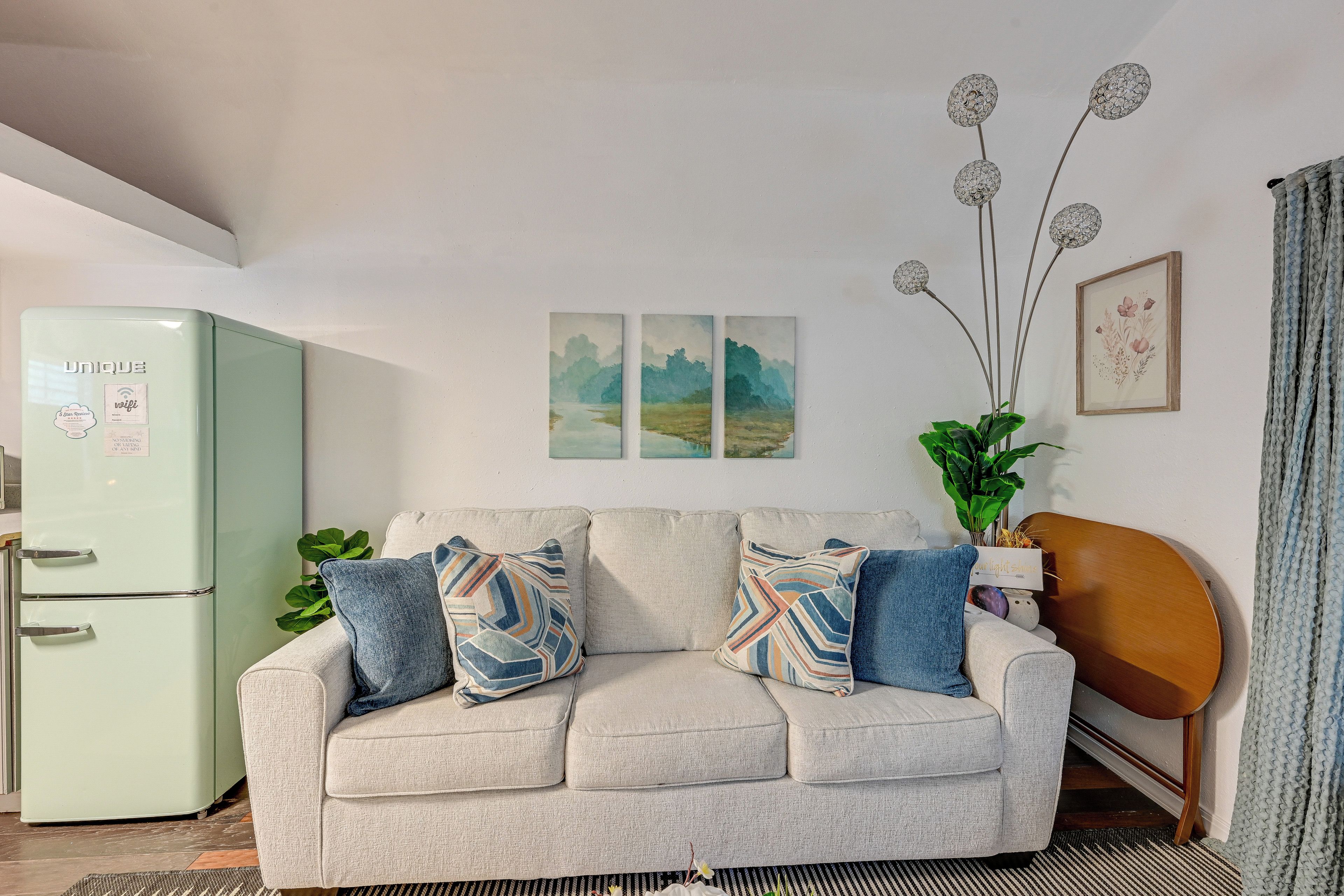 Een woonkamer met een lichtgroene koelkast, een beige bank met gekleurde kussens, een plant en wanddecoratie.
