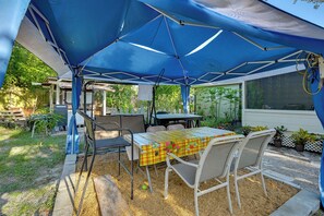 An outdoor patio area with a table and chairs under a blue canopy, surrounded by greenery and a wooden fence.
