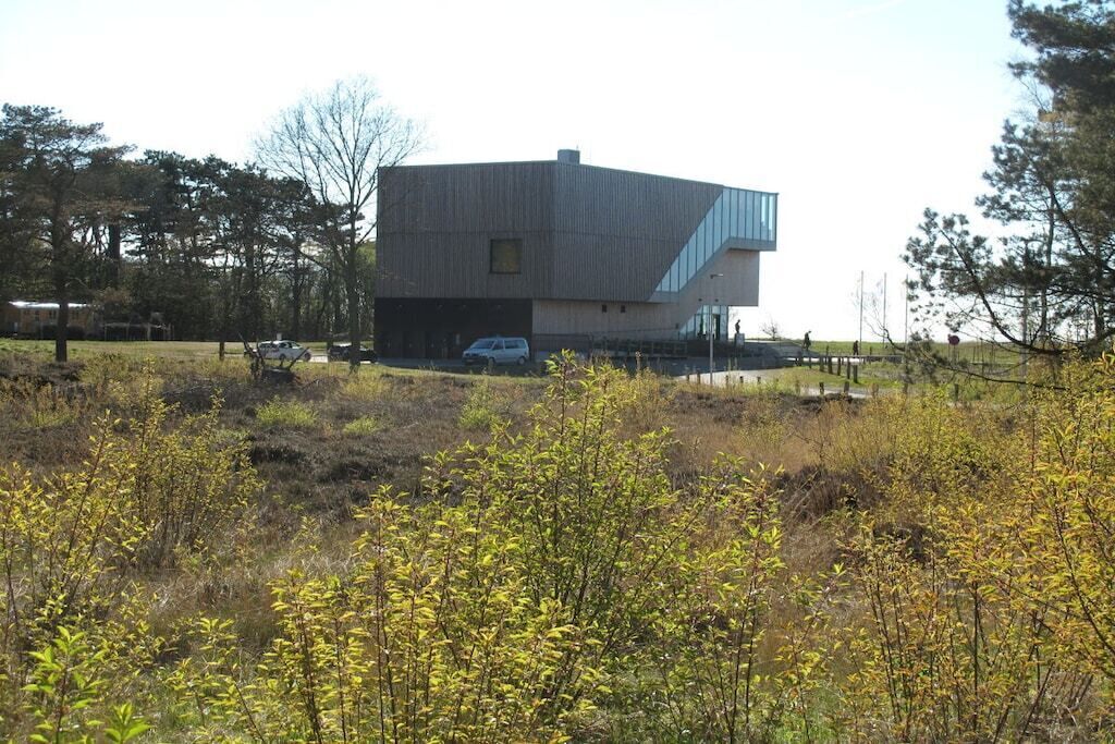 Een modern gebouw met een plat dak en een schuin uitbouw, omgeven door een natuurlijk landschap met weinig begroeiing.
