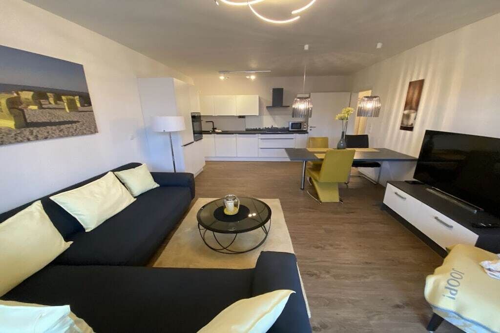 Un salon moderne avec un canapé, une table basse et un téléviseur. On aperçoit, au fond, un coin cuisine avec des placards et un four à micro-ondes.