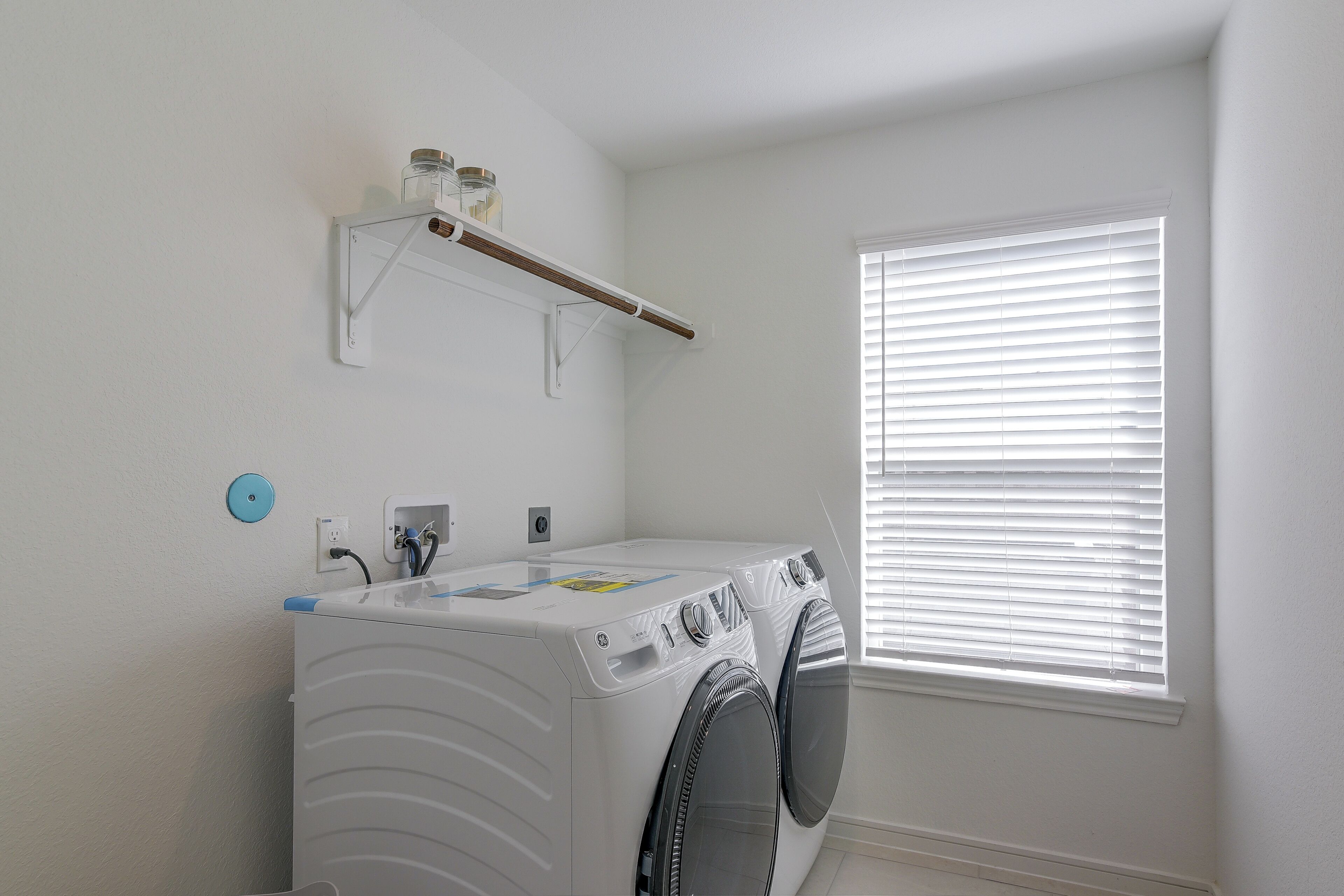 A laundry room with a front-loading washer and dryer, a shelf above the washer, a window with blinds, and a power outlet on the wall.