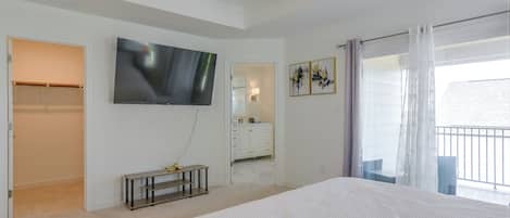 A bedroom with a large bed, a television mounted on the wall, a vanity with a mirror, and a balcony with curtains.
