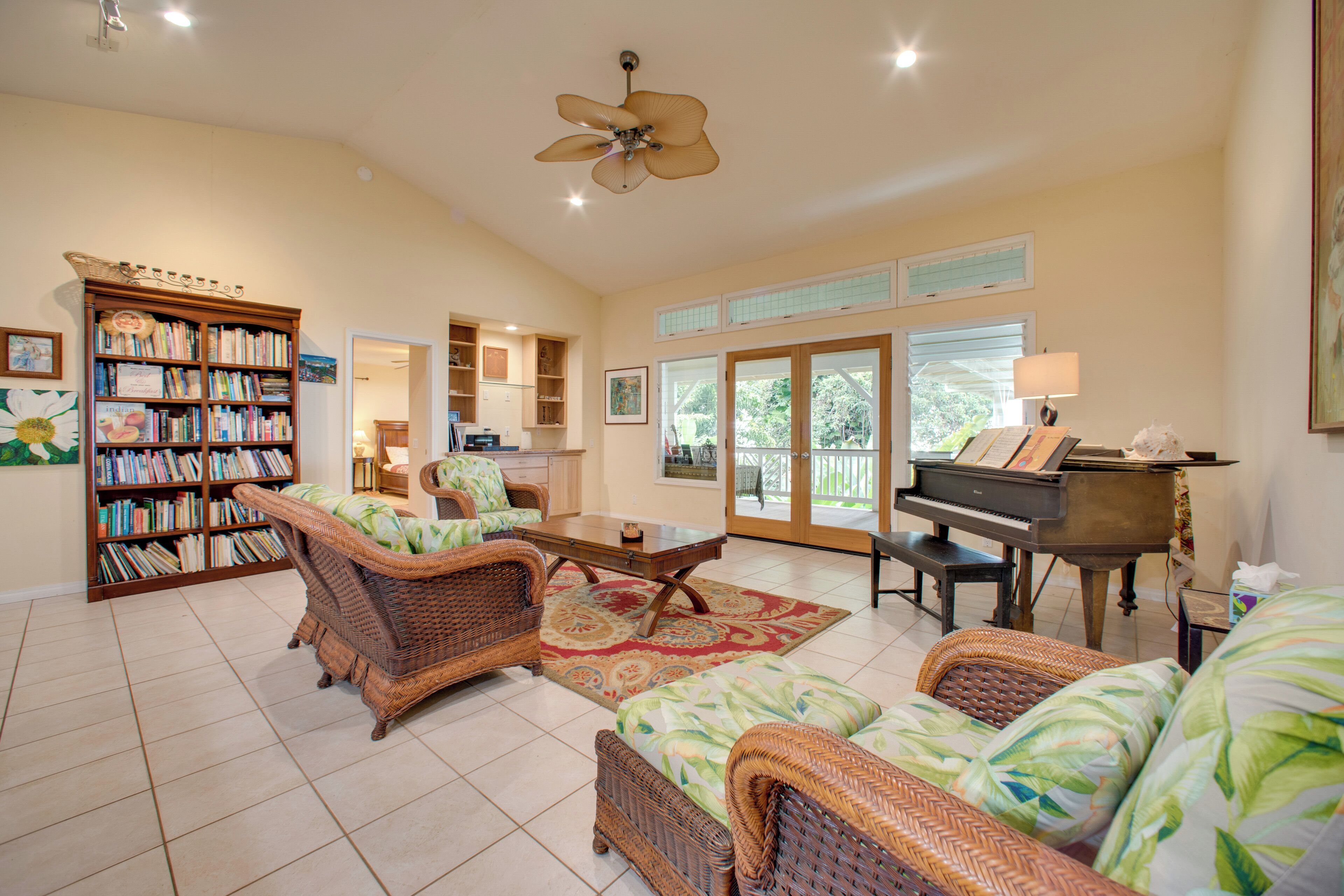 A spacious living room with a piano, wicker furniture, a bookshelf, and a large window.