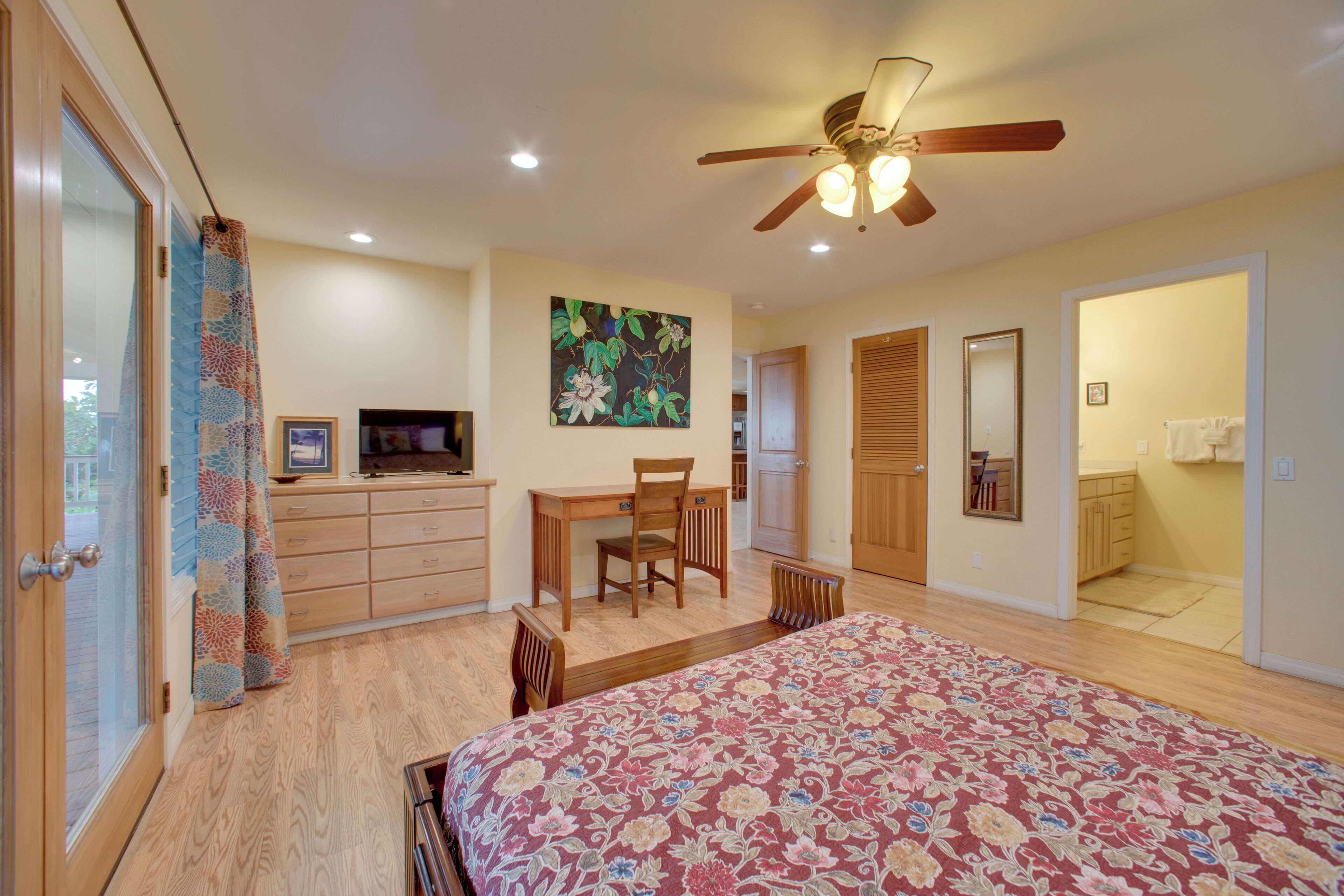 A bedroom with a bed, a desk, a chair, a ceiling fan, and a large window.