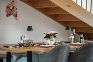 A dining area with a wooden table set for two, featuring a vase with pink flowers, a bottle, and a candle.