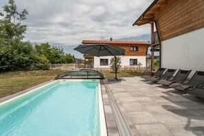 A swimming pool with a sun umbrella, lounge chairs, and a wooden house with a tiled roof.