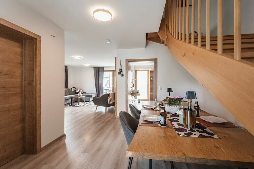 A modern interior with a wooden staircase, a dining area with a table set for two, and a living room in the background.