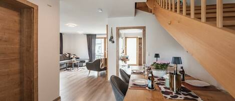 Un interior moderno con una escalera de madera, una zona de comedor con mesa para dos, y una sala de estar al fondo.