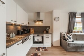 A compact kitchen with white cabinets, a built-in oven, and a stove. There is a grey sofa with a cow print throw pillow and a clock on the wall.