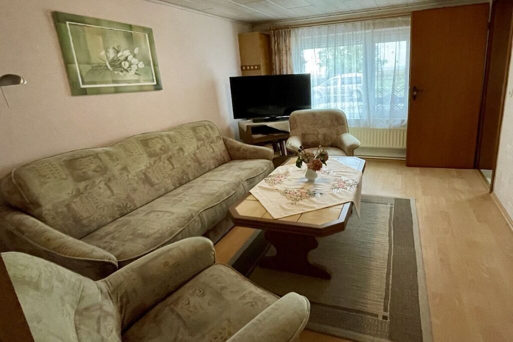 Ein Wohnzimmer mit einer Couch, zwei Sesseln, einem Couchtisch mit einer blumigen Tischdecke, einem Flachbildfernseher und einem Fenster mit Vorhängen.