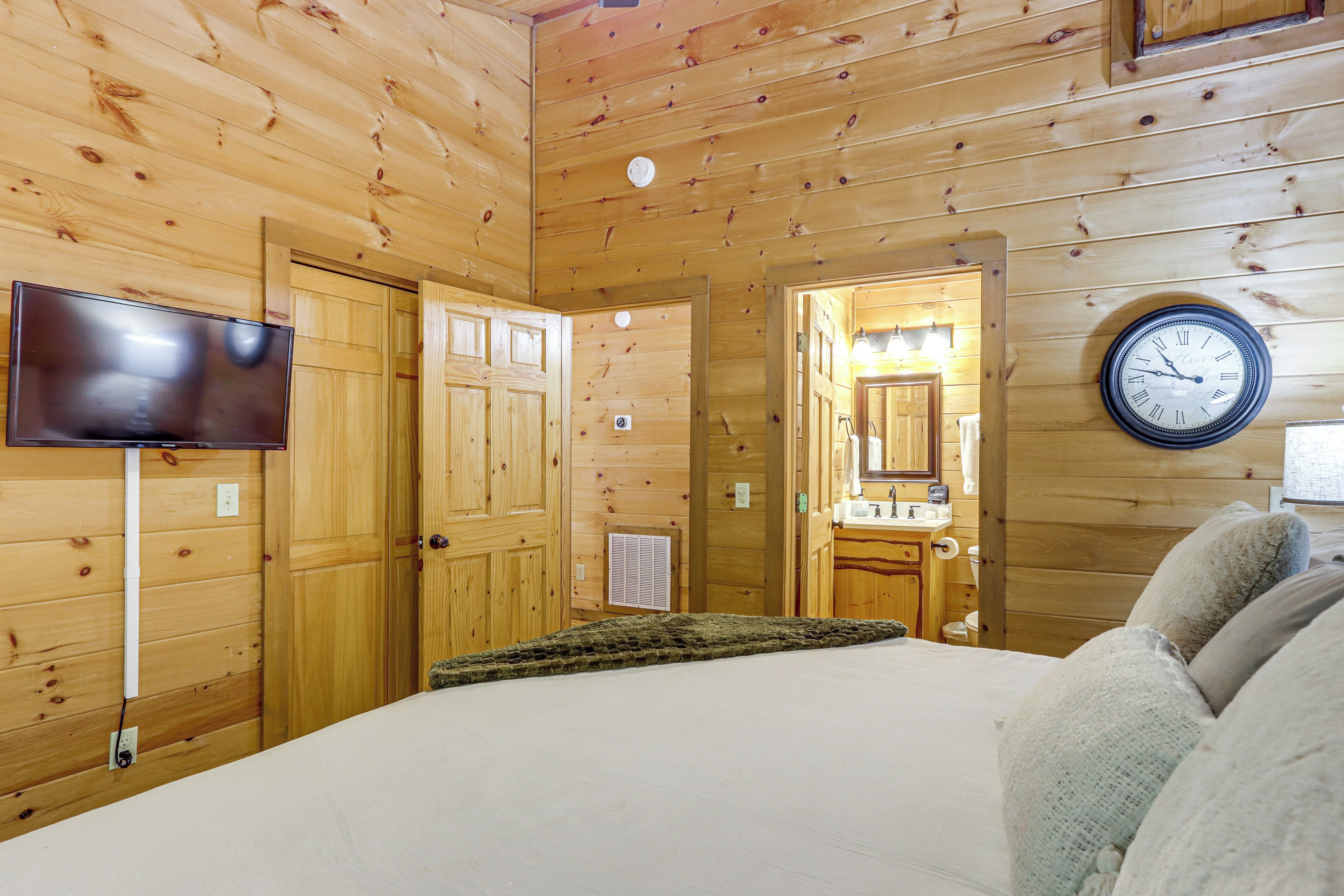 A wooden cabin interior with a bed, a television, a clock, and a bathroom visible through an open door.