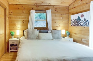 Un dormitorio de cabaña de madera con una cama, dos mesitas de noche, un ventanal con cortinas y un cuadro en la pared.