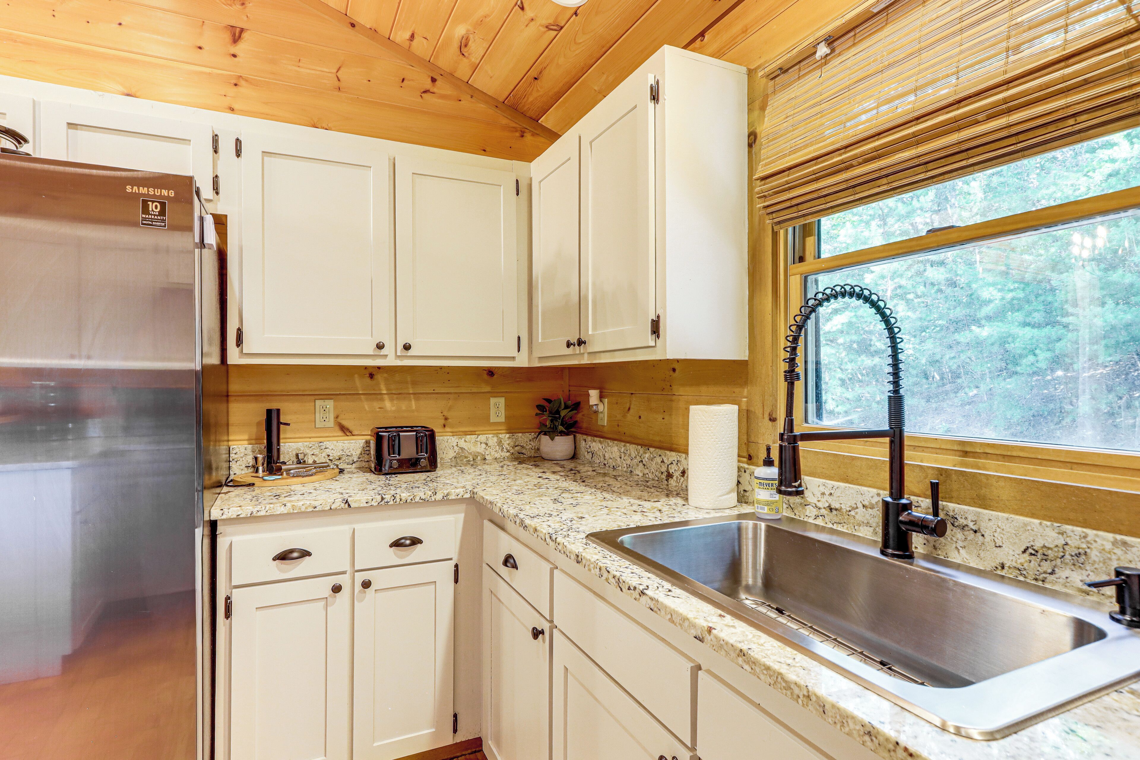 A kitchen with white cabinets, a stainless steel sink, a toaster, and a window with a view of trees.
