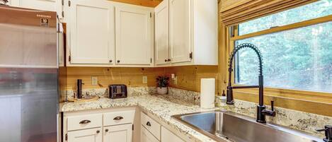 A kitchen with white cabinets, a stainless steel sink, a toaster, and a window with a view of trees.