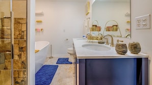 A modern bathroom with a stone wall, a large mirror, a white sink, a gold-toned vanity, and a blue bath mat. - Home Gym & Pool Access! North Raleigh Retreat (Raleigh)