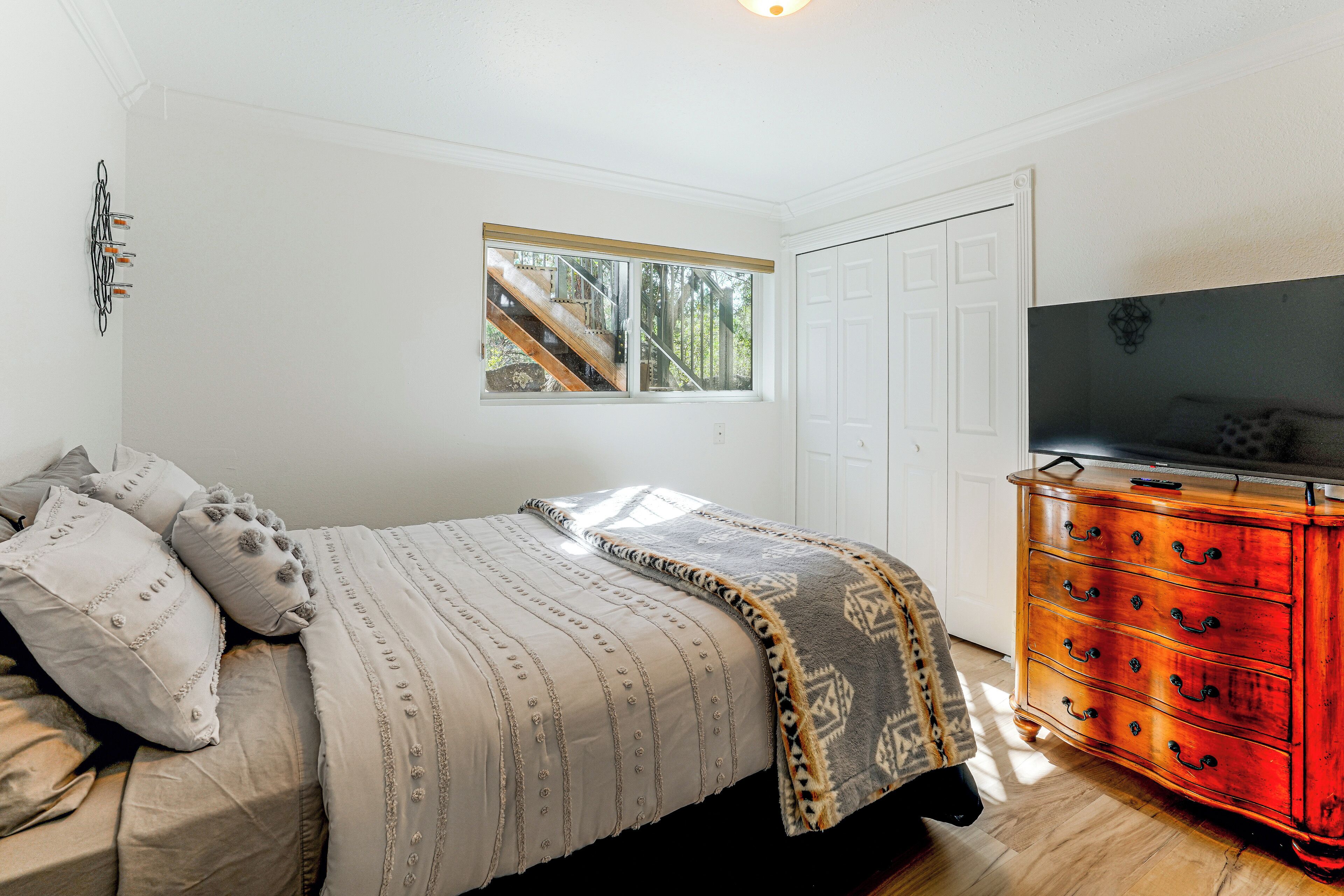 A bedroom with a bed, a dresser, a television, and a window with a view of a staircase.