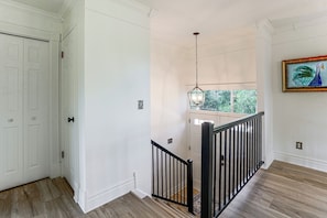 Una escalera con barandilla negra lleva a una plataforma, una puerta blanca a la izquierda y una ventana con vista a follaje.