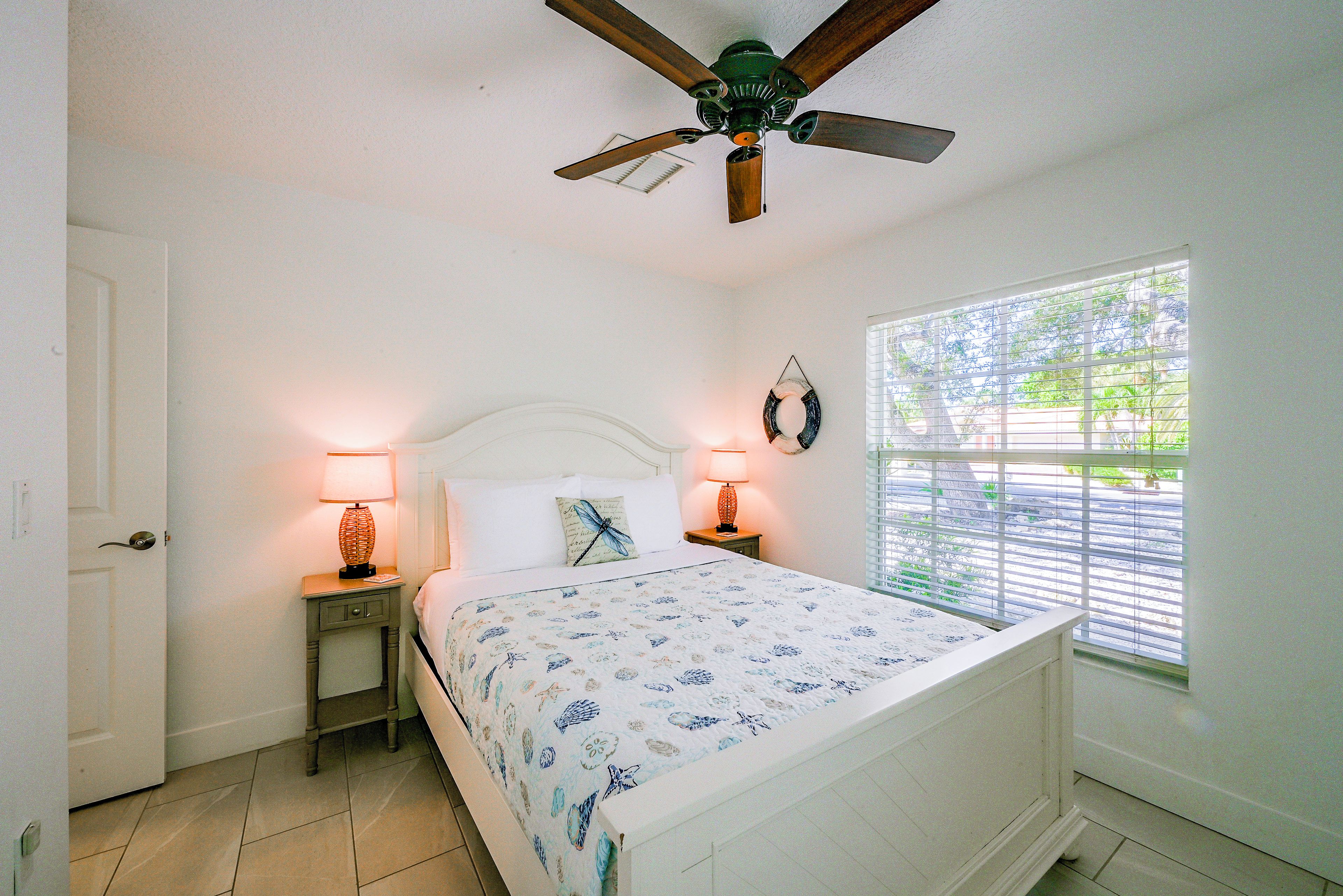 A bedroom with a bed, two bedside lamps, a ceiling fan, a window with blinds, and a door.
