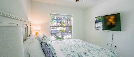 Un dormitorio con cama, ventilador de techo, televisión montada en la pared y ventana con persianas.