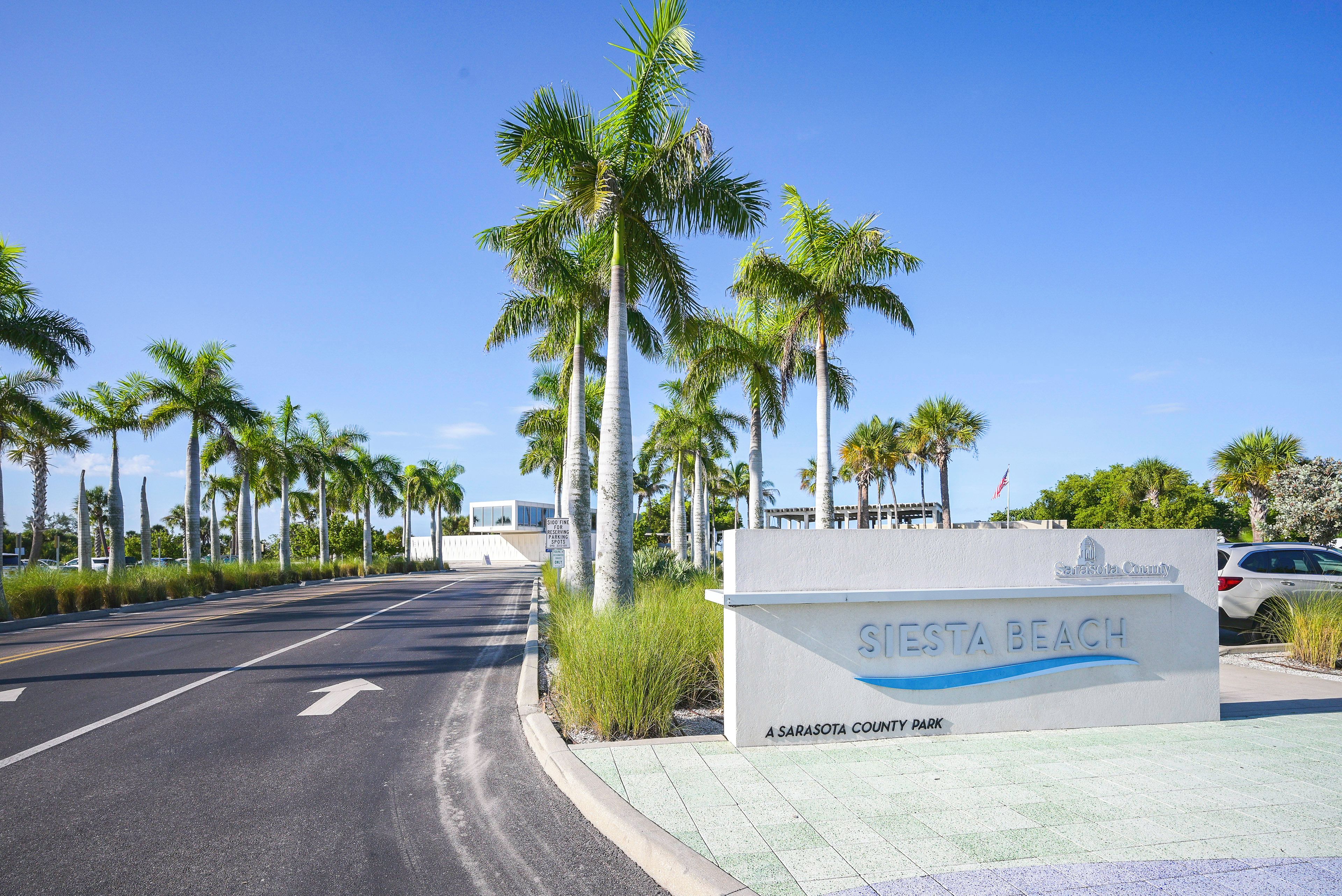 A road leading to a sign for 'SIESTA BEACH' with a clear sky and palm trees.