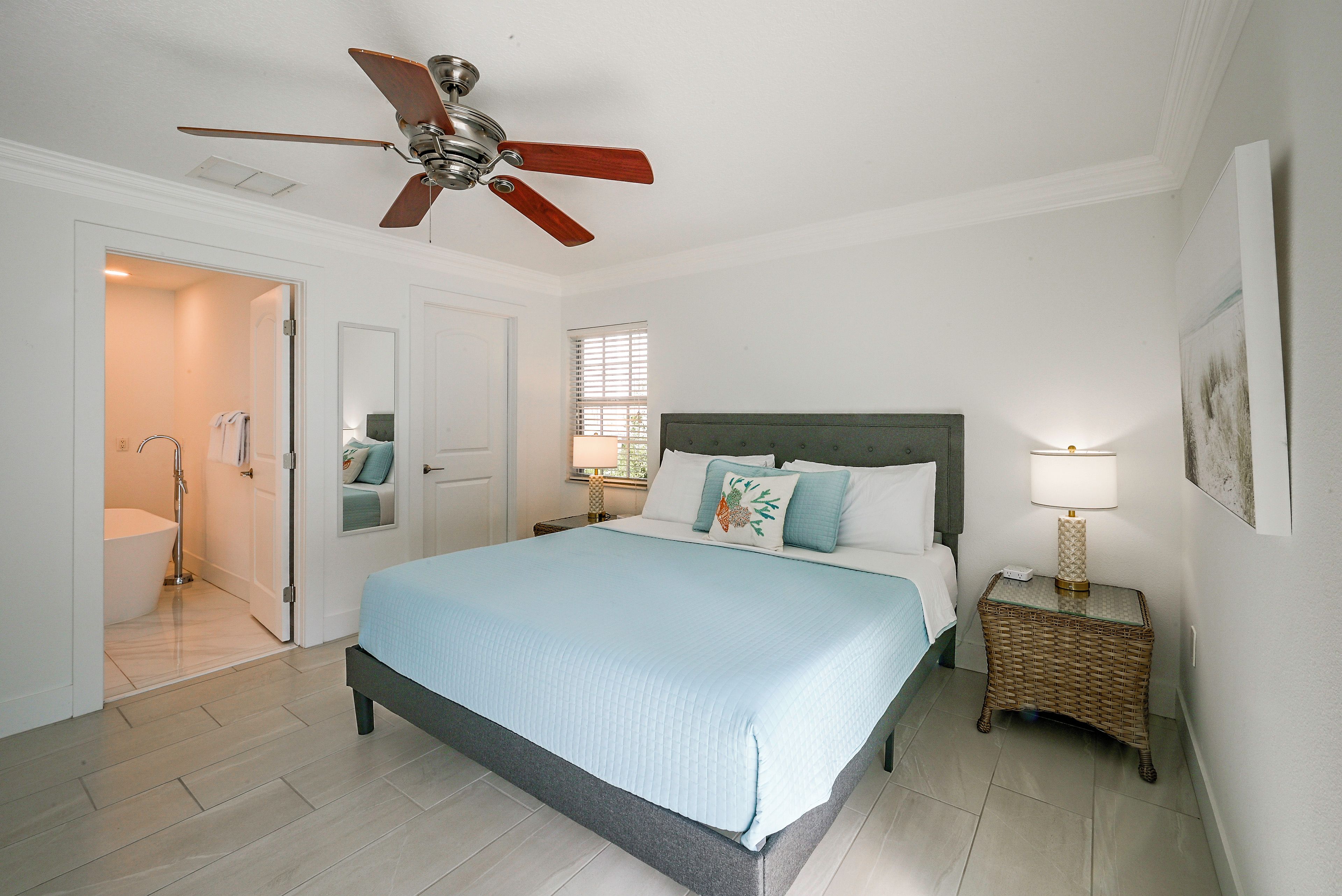 A bedroom with a bed, a bedside table, a lamp, a ceiling fan, a window with blinds, and a door leading to a bathroom.