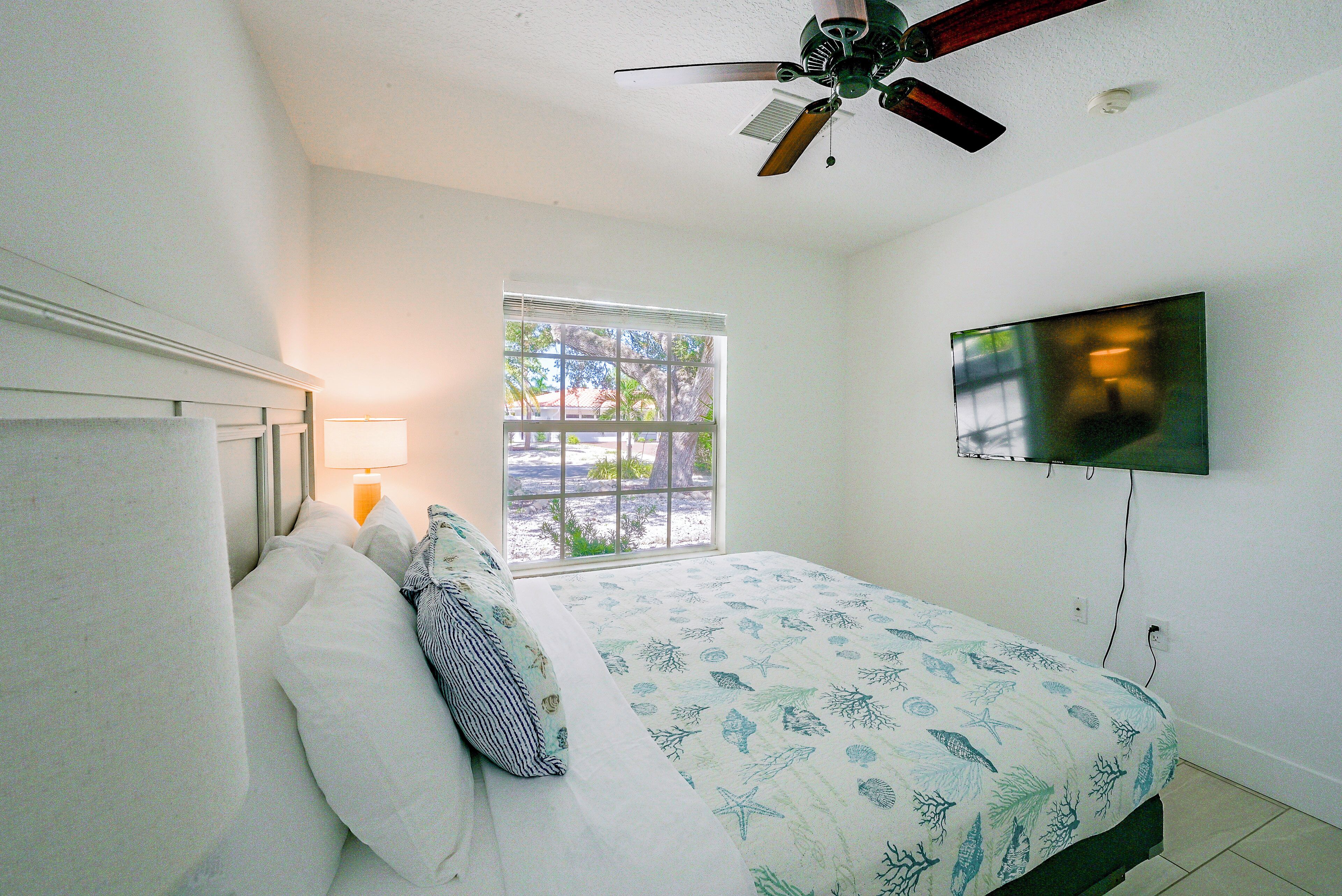 A bedroom with a bed, a ceiling fan, a TV mounted on the wall, and a window with blinds.