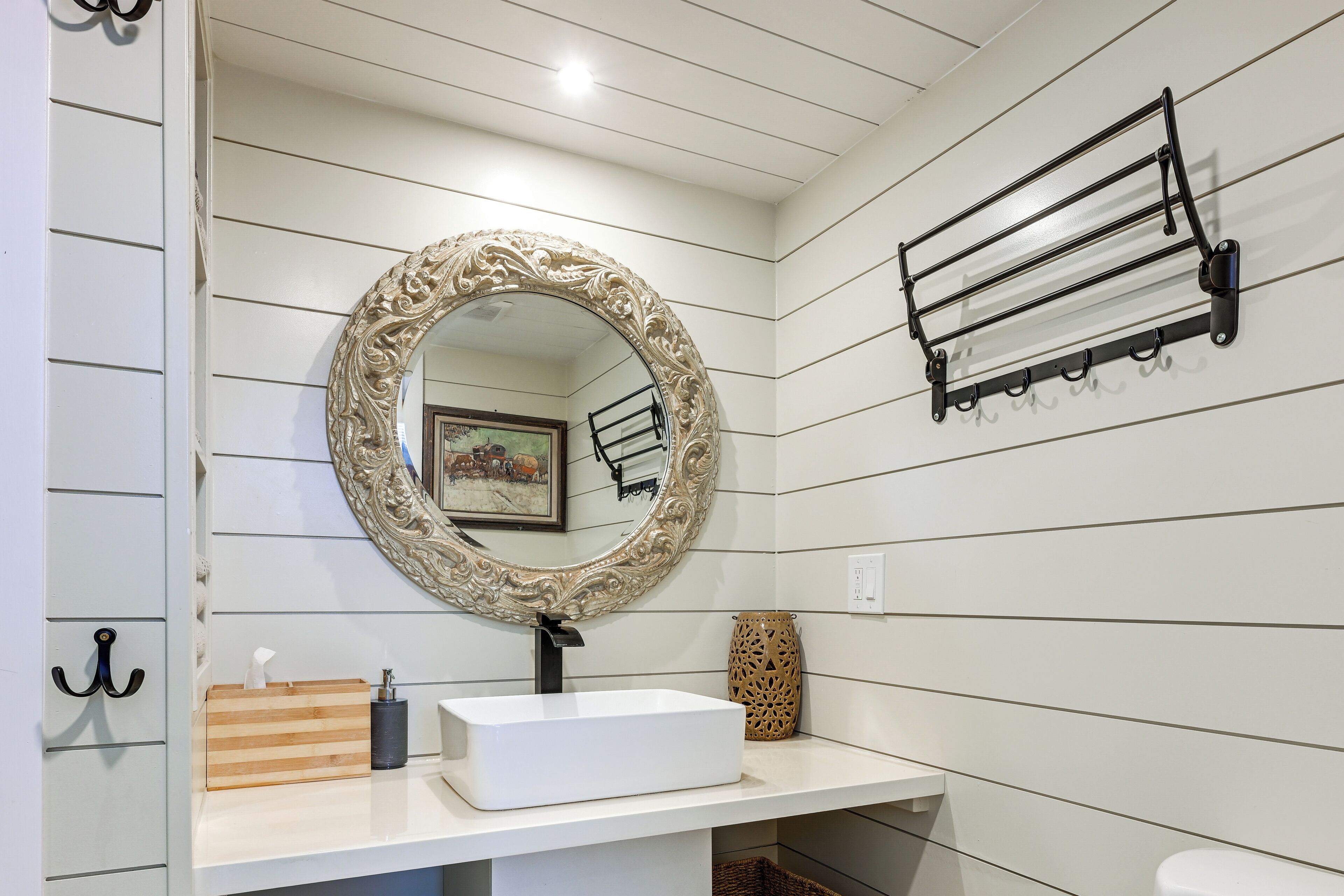 A bathroom with a white vanity, a large ornate mirror, a towel rack, and a framed picture on the wall.