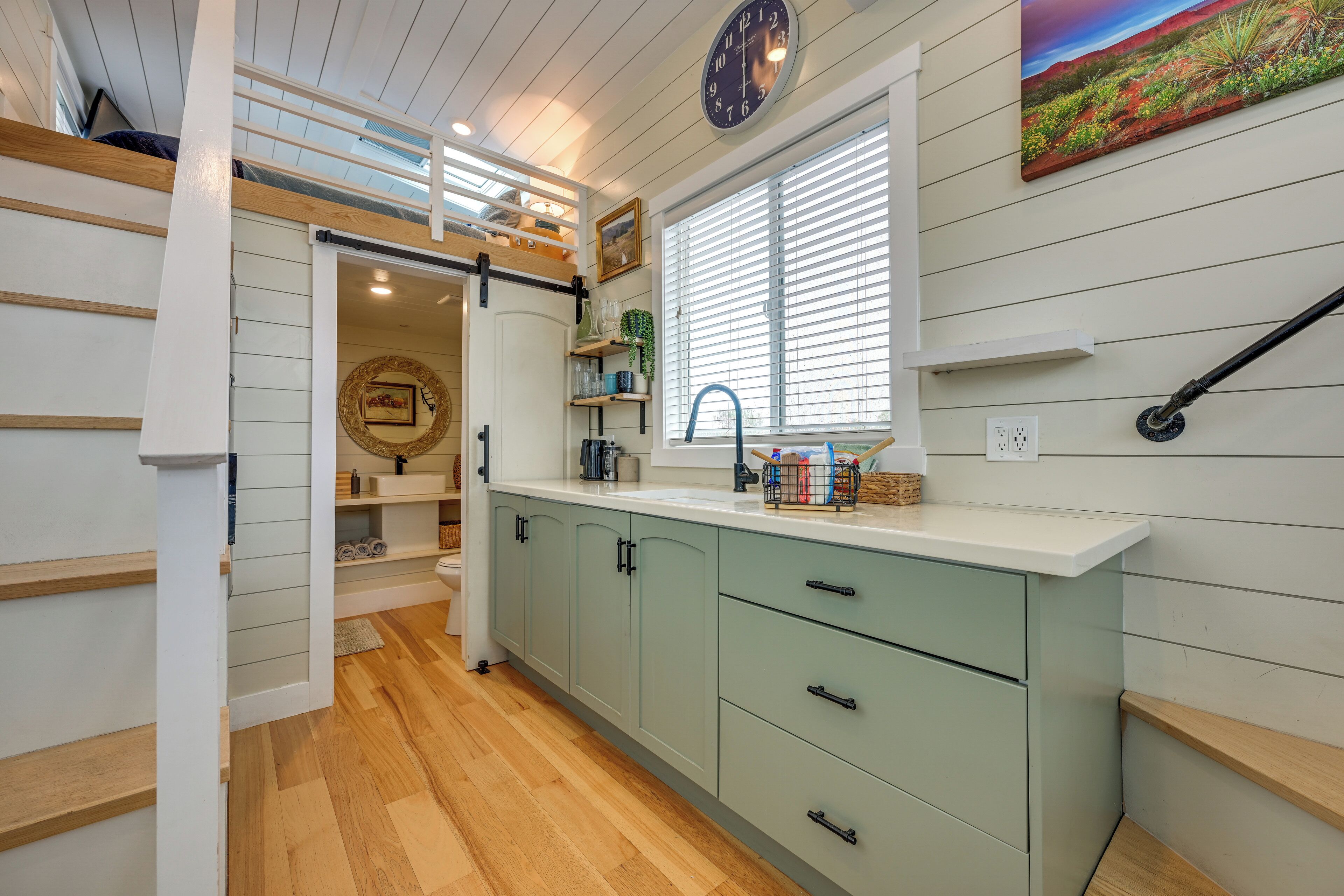 A kitchen with a light green cabinet, a window with blinds, a clock on the wall, and a painting above the window.